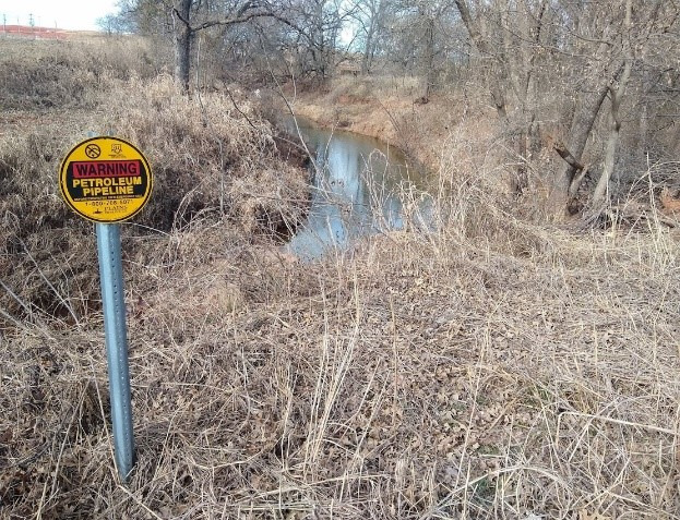 A creek in Logan County, OK where Stephenson played as a child. A recently built pipeline warning sign stands beside the creek and a new drilling site looms in the distance